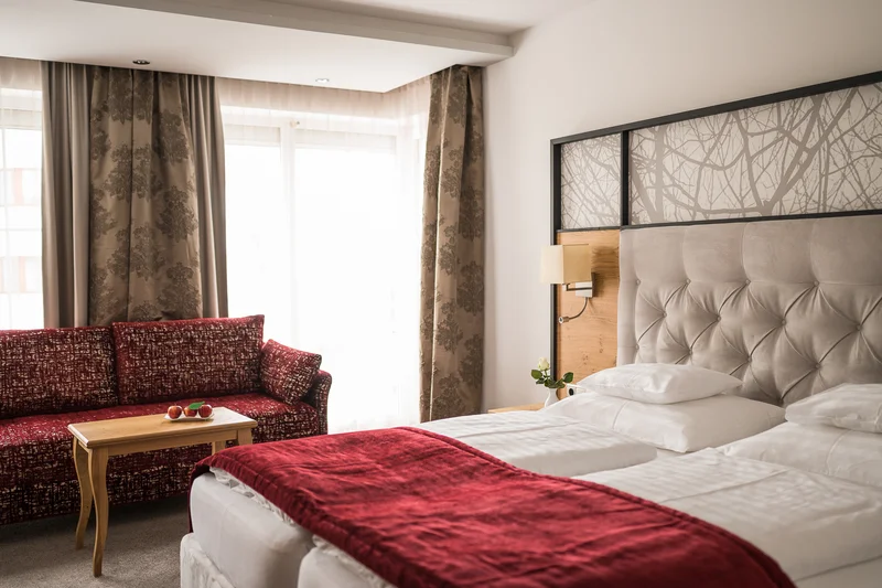 Hell eingerichtetes Hotelzimmer mit Doppelbett, roter Decke und Sofa am Fenster
