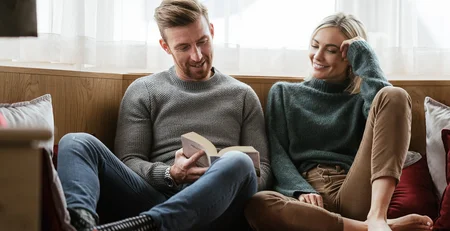 Die besten Referenzen für unser Hotelmarketing Paar sitzt gemütlich auf Sofa, Mann liest Buch, Frau hört lächelnd zu