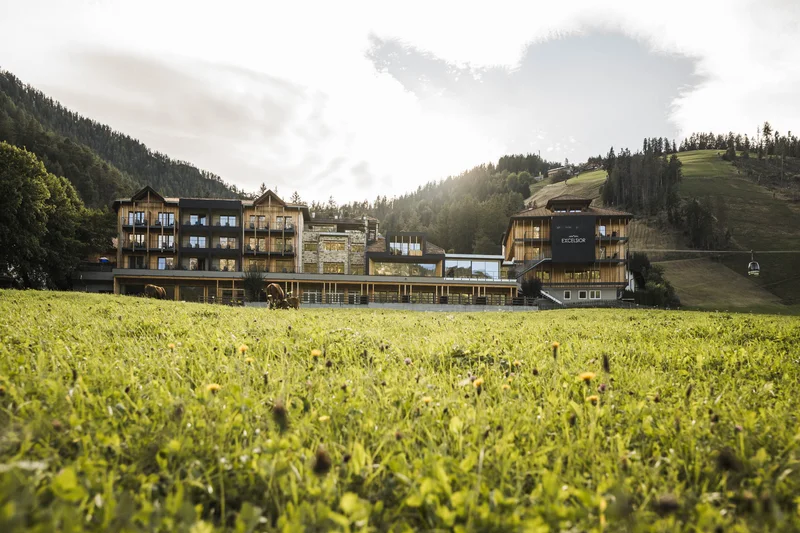 Follow the call of the mountains to St. Vigil Hotel Excelsior with meadow, cable car, and forested hillside in the background