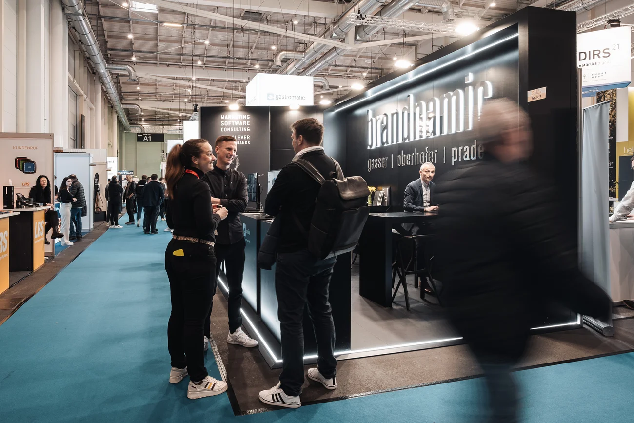 Menschen unterhalten sich an einem Messestand mit beleuchtetem brandteam Schriftzug