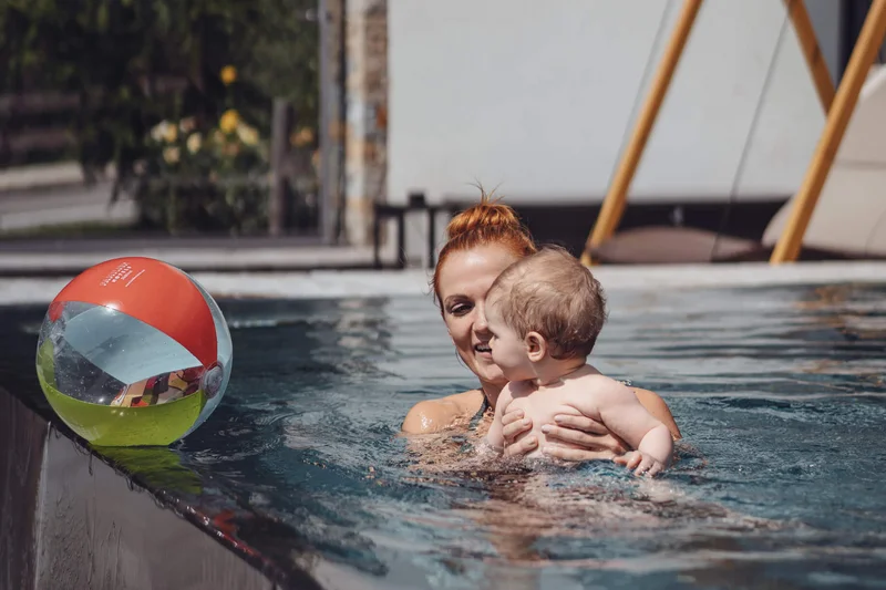 Die Familienhotels Südtirol – branded by Brandnamic Frau hält Kleinkind im Pool mit buntem Strandball am Beckenrand