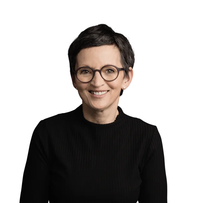 Helga Karbon Frau mit kurzen dunklen Haaren, runder Brille und schwarzem Pullover lächelnd