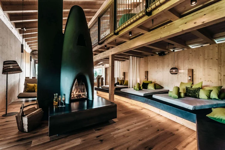 Der rote Faden im Hotel: Warum Erlebnisse konsistent sein müssen Holz-Lounge mit großem schwarzem Kamin, gemütlichen Sitzbänken und grünen Kissen