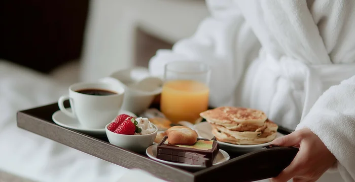 Person in Bademantel hält Frühstückstablett mit Kaffee, Saft, Pfannkuchen und Obst