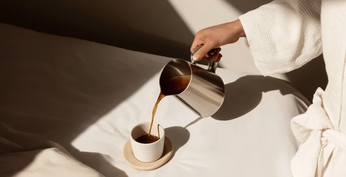 Hand gießt Kaffee aus Metallkanne in Tasse auf Bett mit Tageslicht
