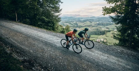 Two cyclists riding on a gravel road in a hilly, forested landscape