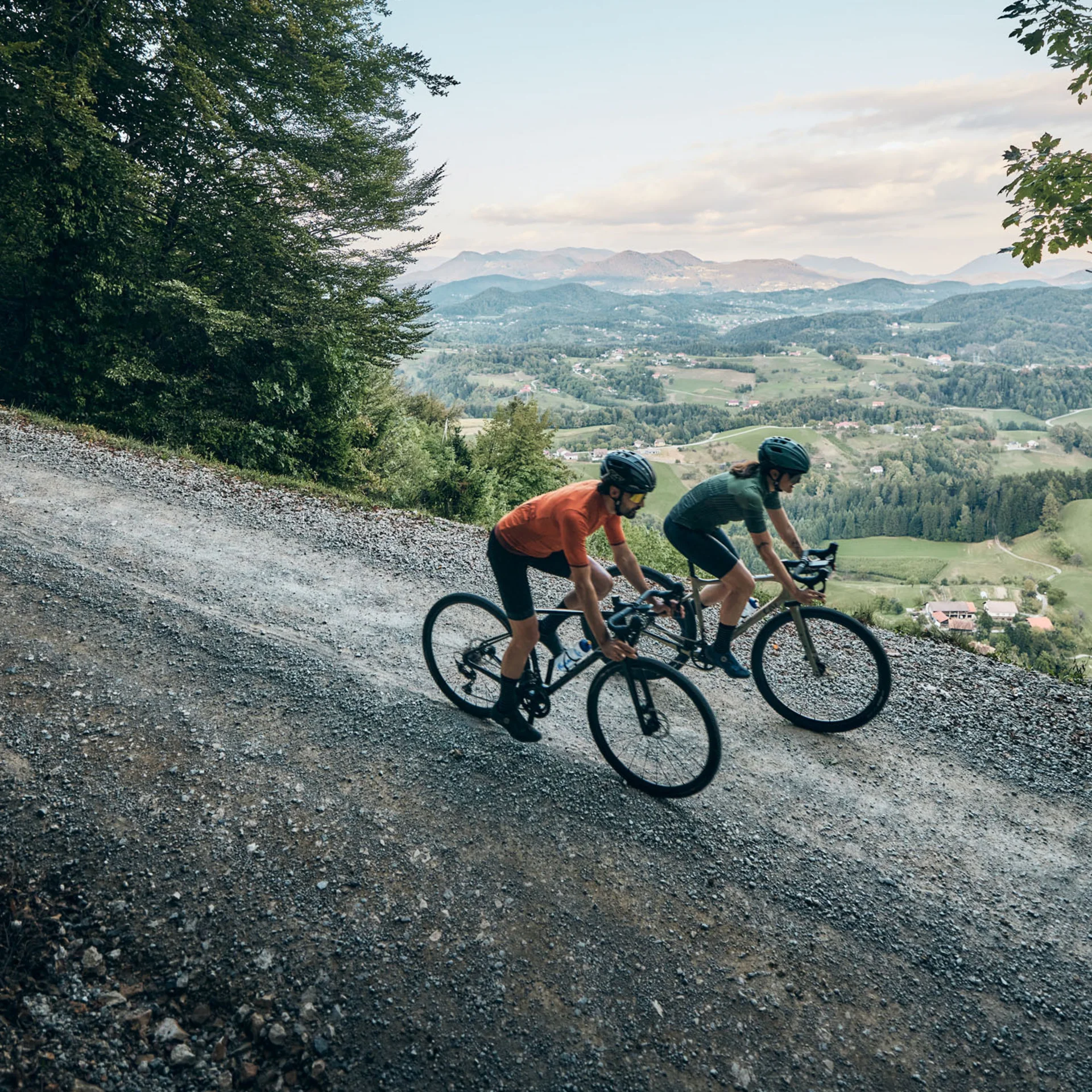 Zwei Radfahrer fahren auf einem Schotterweg in hügeliger, bewaldeter Landschaft