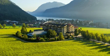 Hotel in grünem Tal vor Bergen und See im Sonnenlicht